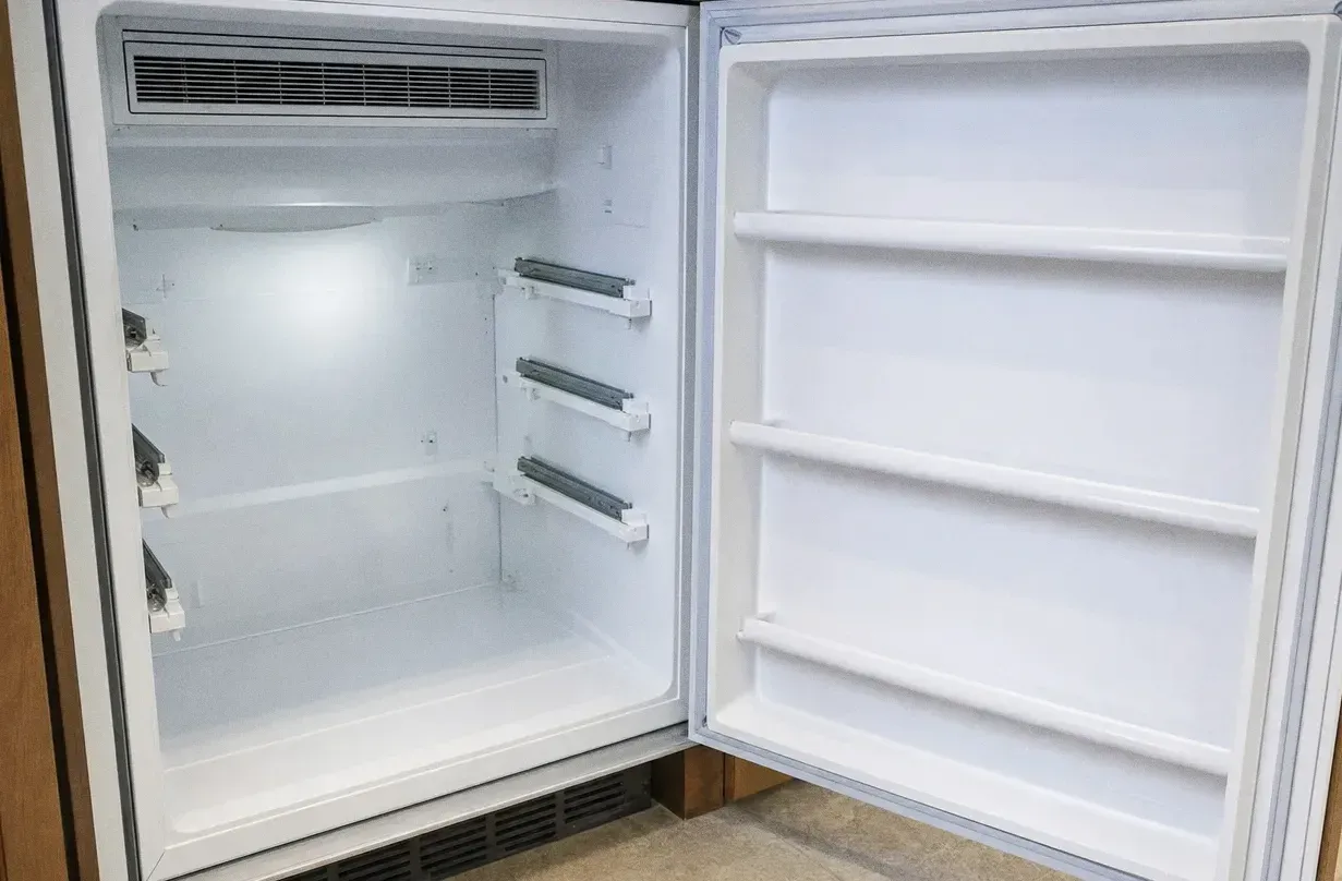 Compact white refrigerator standing open with bare shelves, empty door bins and the vent grille across the top of the cabinet