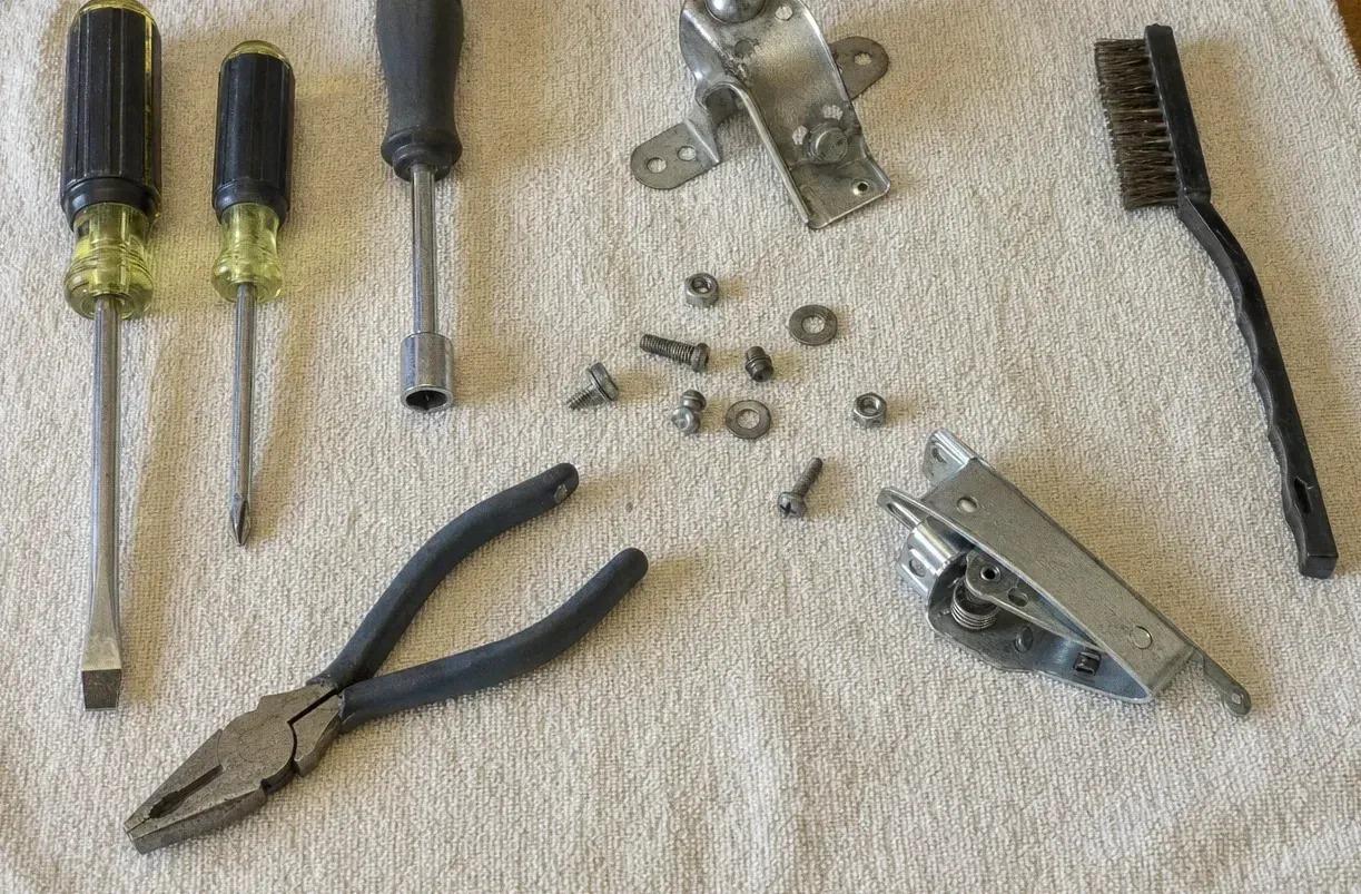 Door latch assembly with its striker laid out beside screwdrivers, a nut driver, lineman’s pliers and a wire brush on a towel